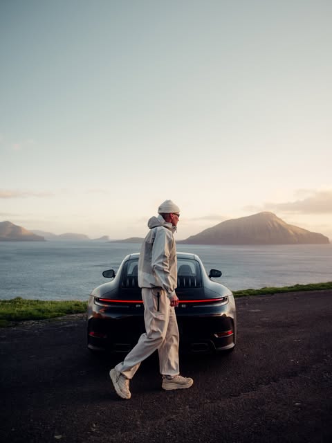 A figure walks past a Porsche 911 on a coastal road at golden hour